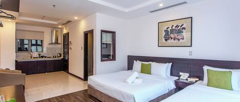 A hotel room with two beds, a small desk, a chair, a dining table, and a kitchen area in the background.