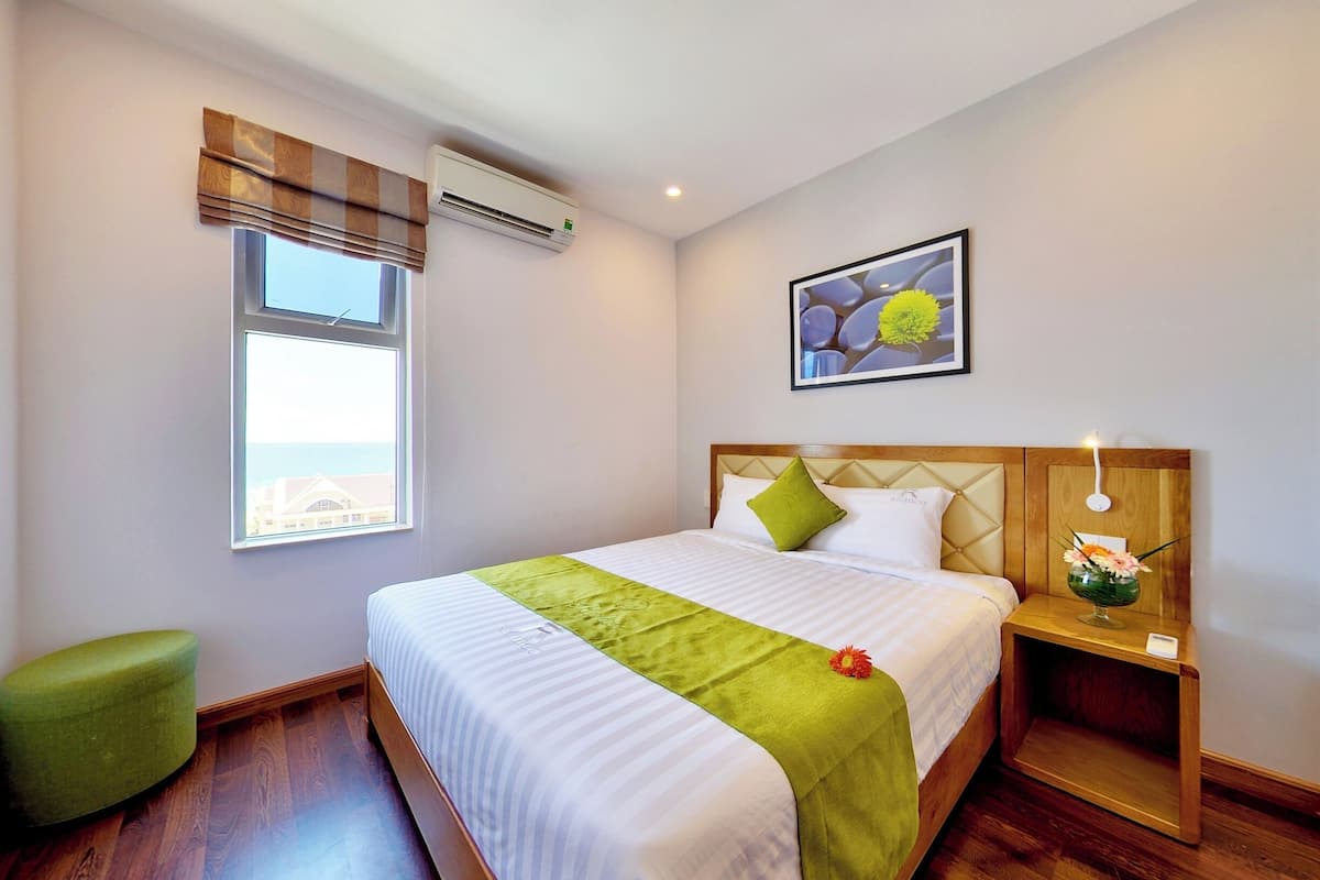 Una cama bien hecha con un plaid verde, una mesita de noche con lámpara y una ventana con persianas.