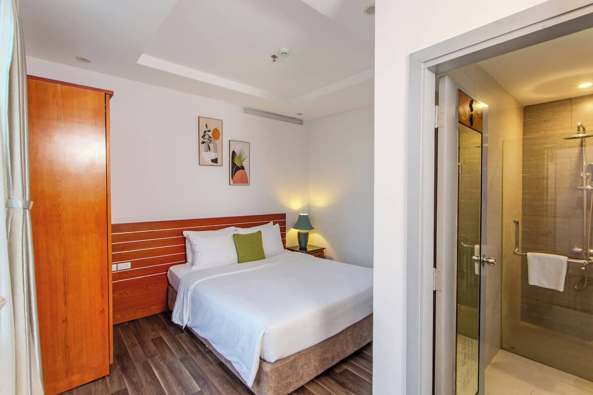 Habitación de hotel con cama de cabecera de madera, mesita de noche con lámpara y cabina de ducha de vidrio.