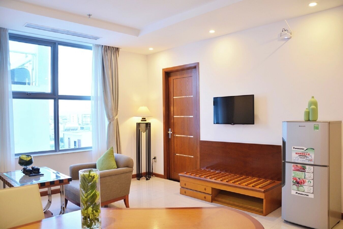 Una camera d'albergo moderna con una panca in legno, un tavolo da pranzo con sedie, una lampada da terra, una TV a schermo piatto e un frigorifero.