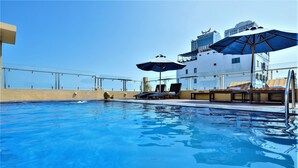 Une piscine avec des chaises longues et des parasols, un ciel dégagé et, au fond, un bâtiment sur lequel on peut lire l’inscription « Sunny ».