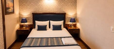 Una habitación de hotel con una cama grande, dos mesitas de noche con lámparas, un papel tapiz estampado y un cuadro enmarcado en la pared izquierda.