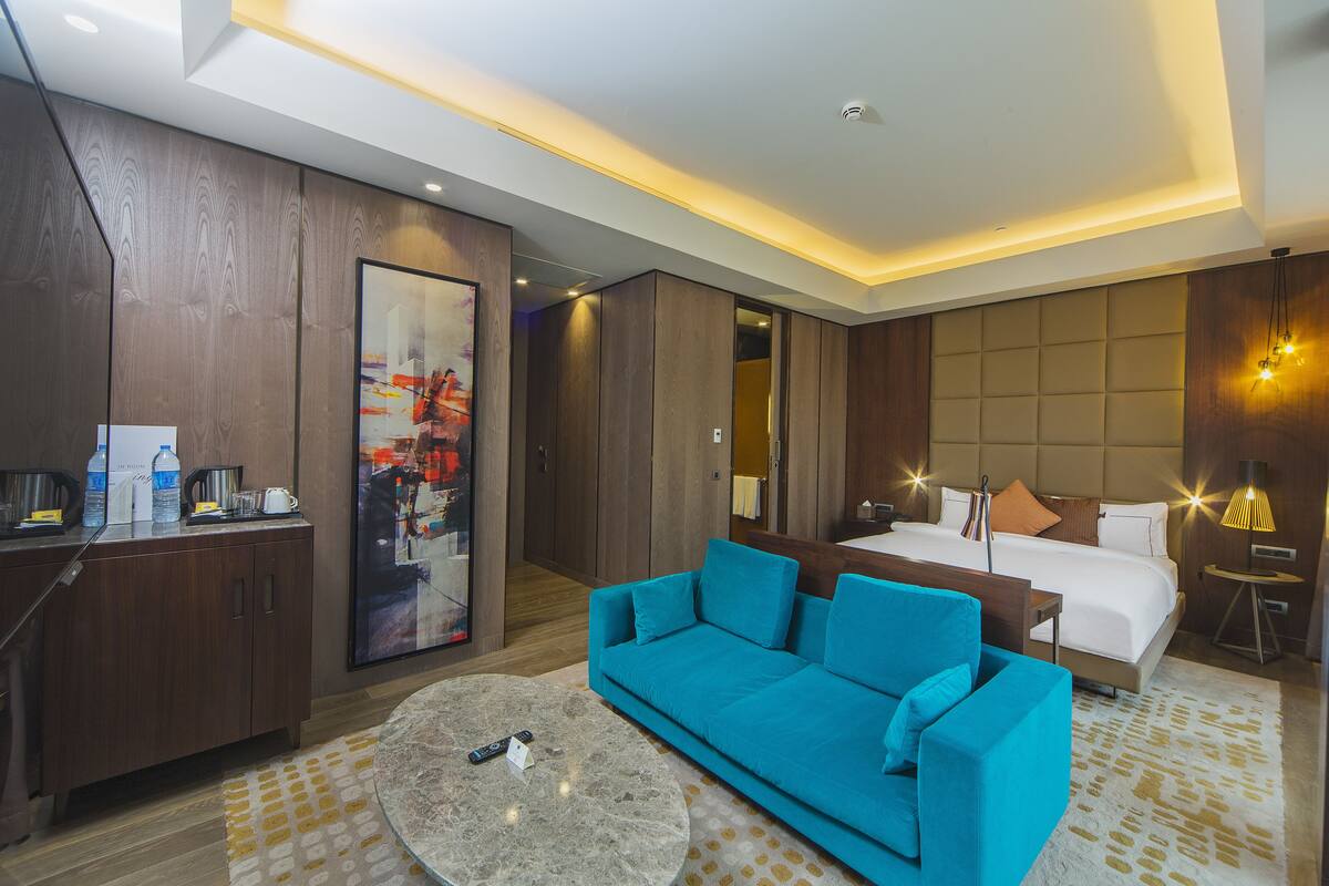 A modern hotel room with a large bed, a blue sofa, a desk with a coffee maker, and a large painting on the wall.