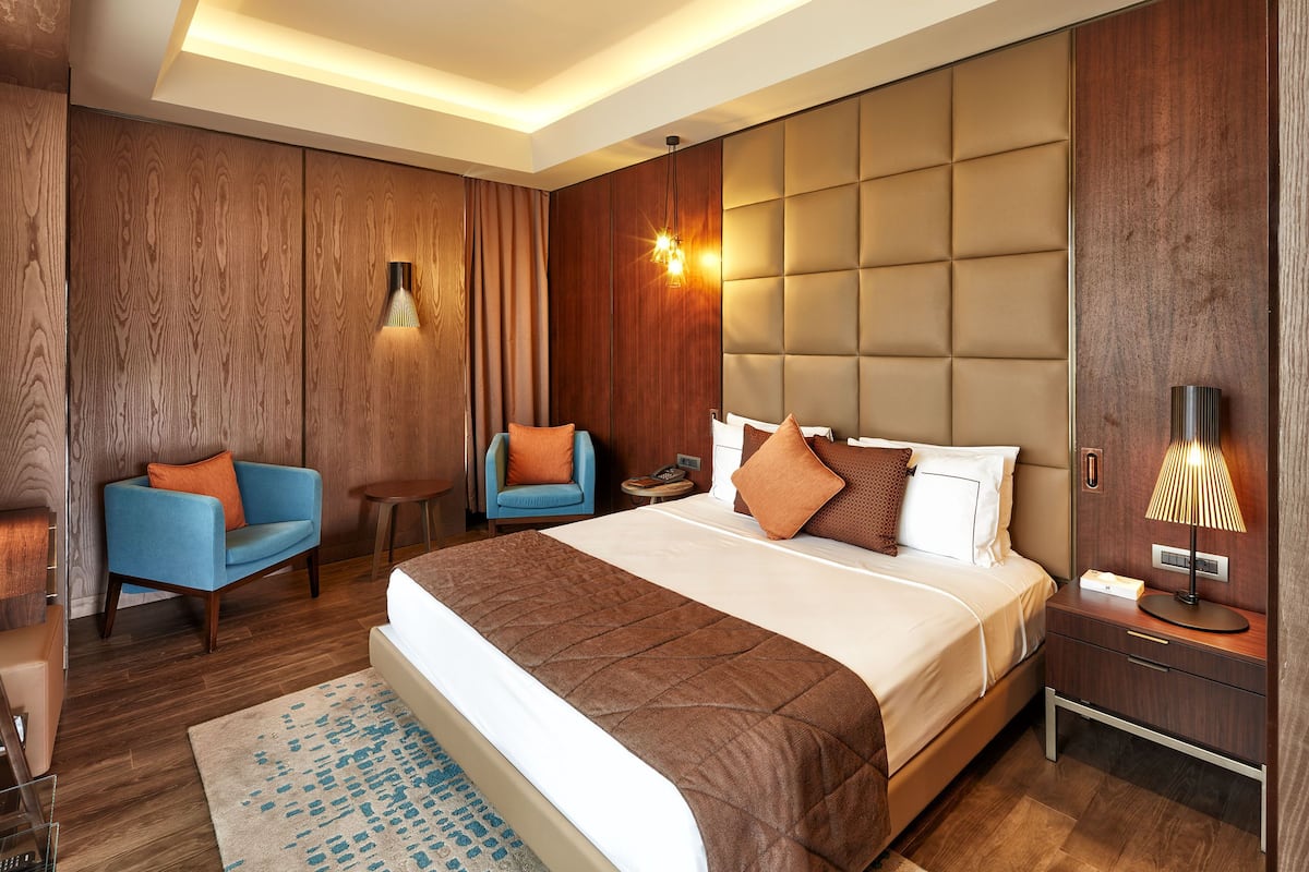 A modern hotel room with a large bed, a seating area with a blue armchair, a wooden accent wall, and a nightstand with a lamp.