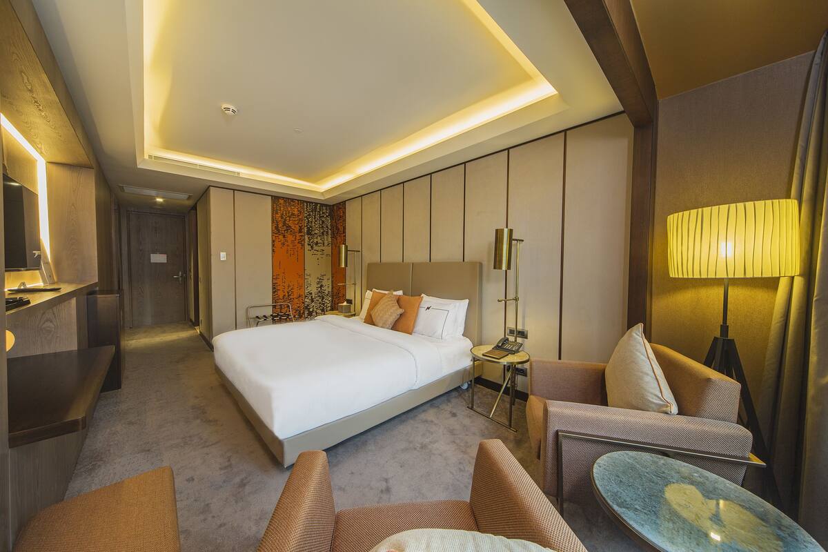A modern hotel room with a large bed, a sofa, a small table, and a desk with a lamp.