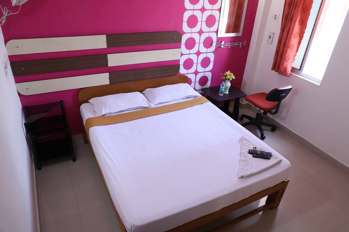 Uma cama bem arrumada com roupa de cama branca, cabeceira de madeira e uma pequena escrivaninha com cadeira.