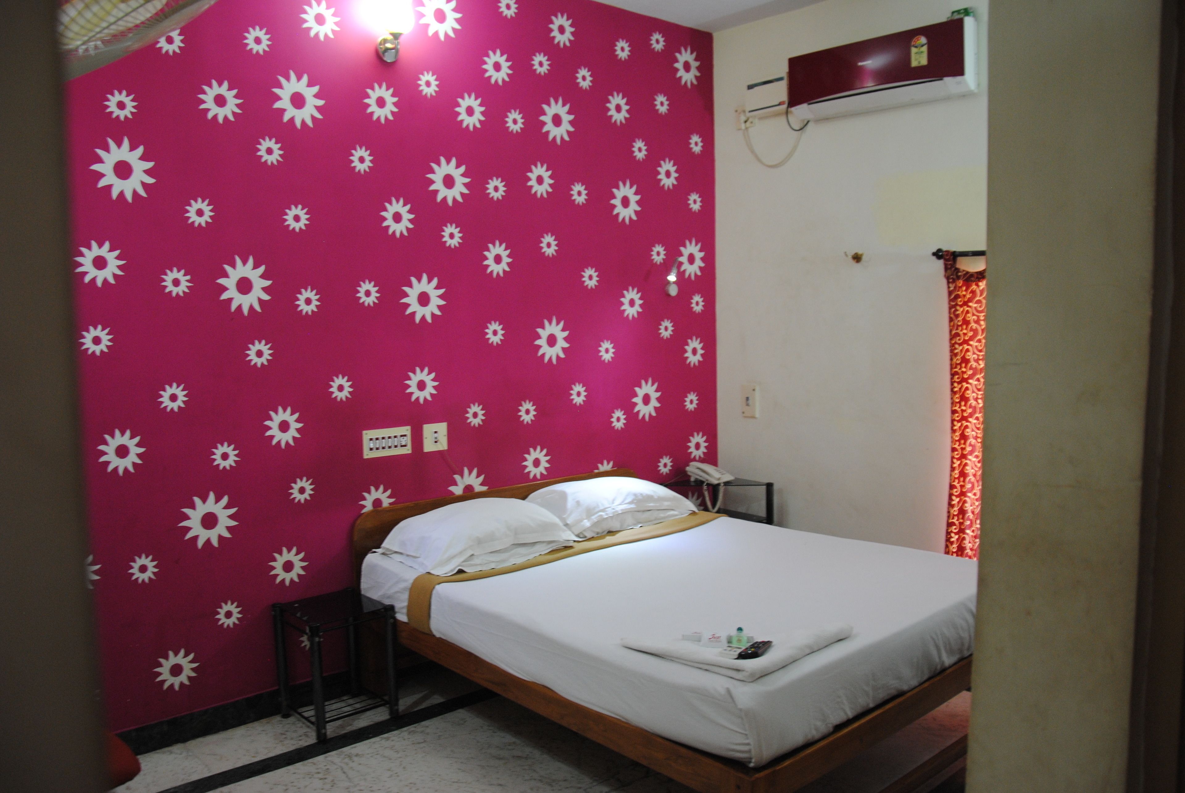 A bed in a room with a pink wall featuring white sun patterns, a wooden headboard, a bedside table, and a red curtain.