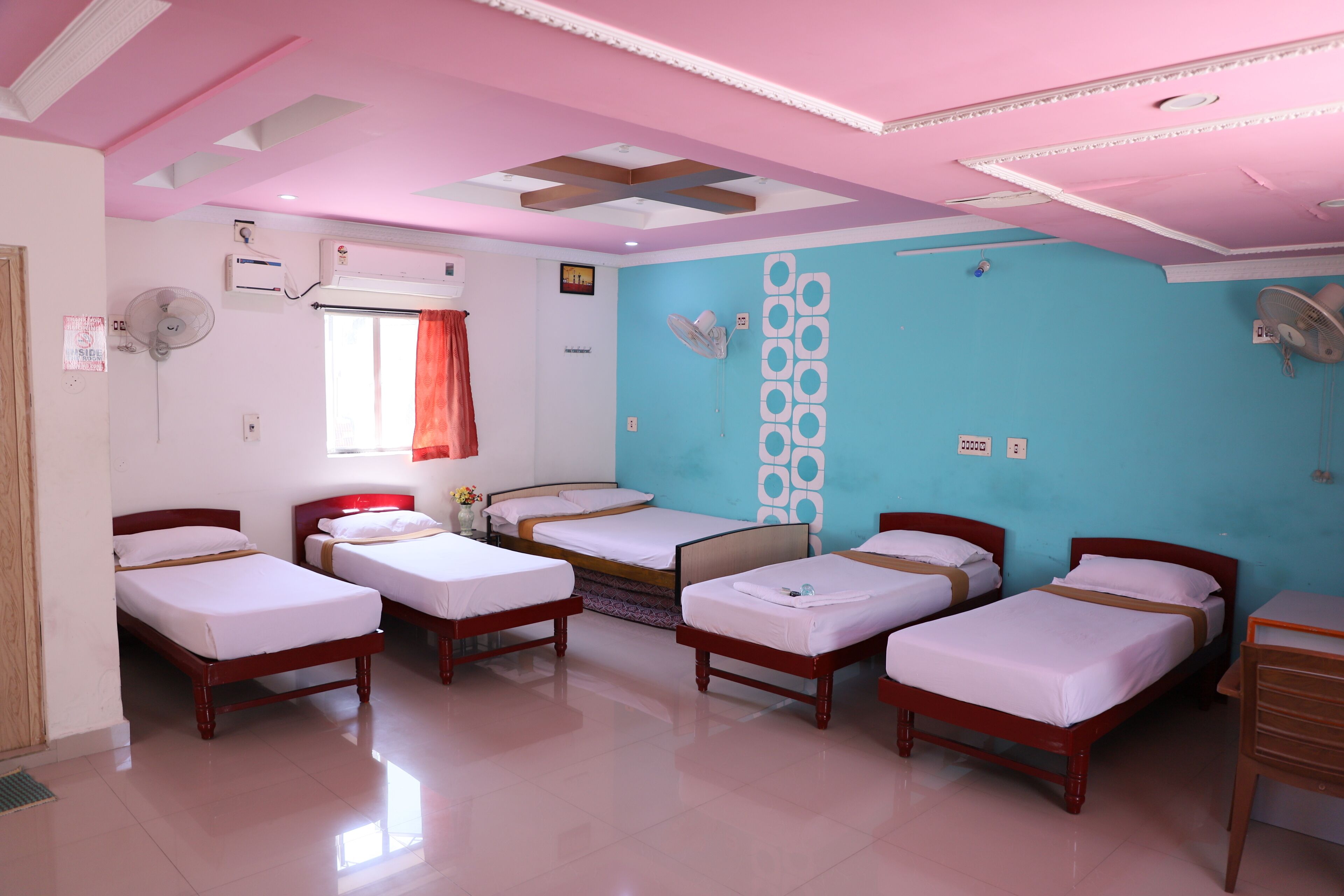 A hotel room with six single beds, a wooden desk, a window with a red curtain, and a blue accent wall with a decorative pattern.