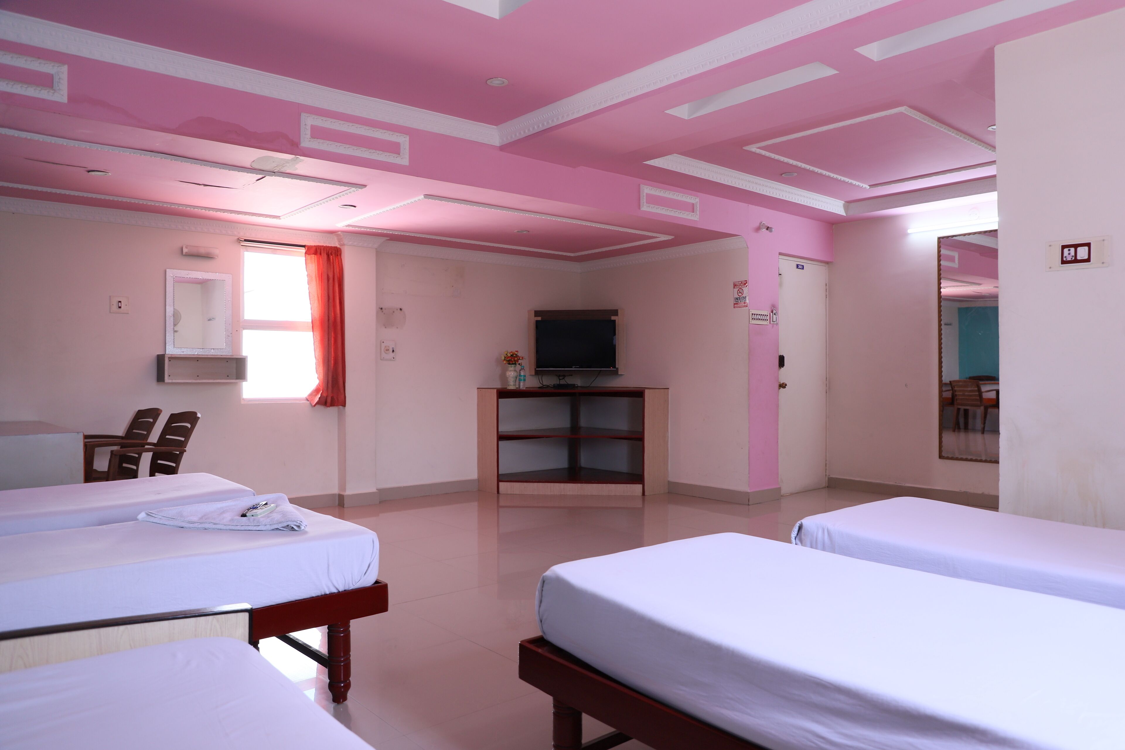 A hotel room with two single beds, a desk with a chair, a television on a stand, and a window with a red curtain.