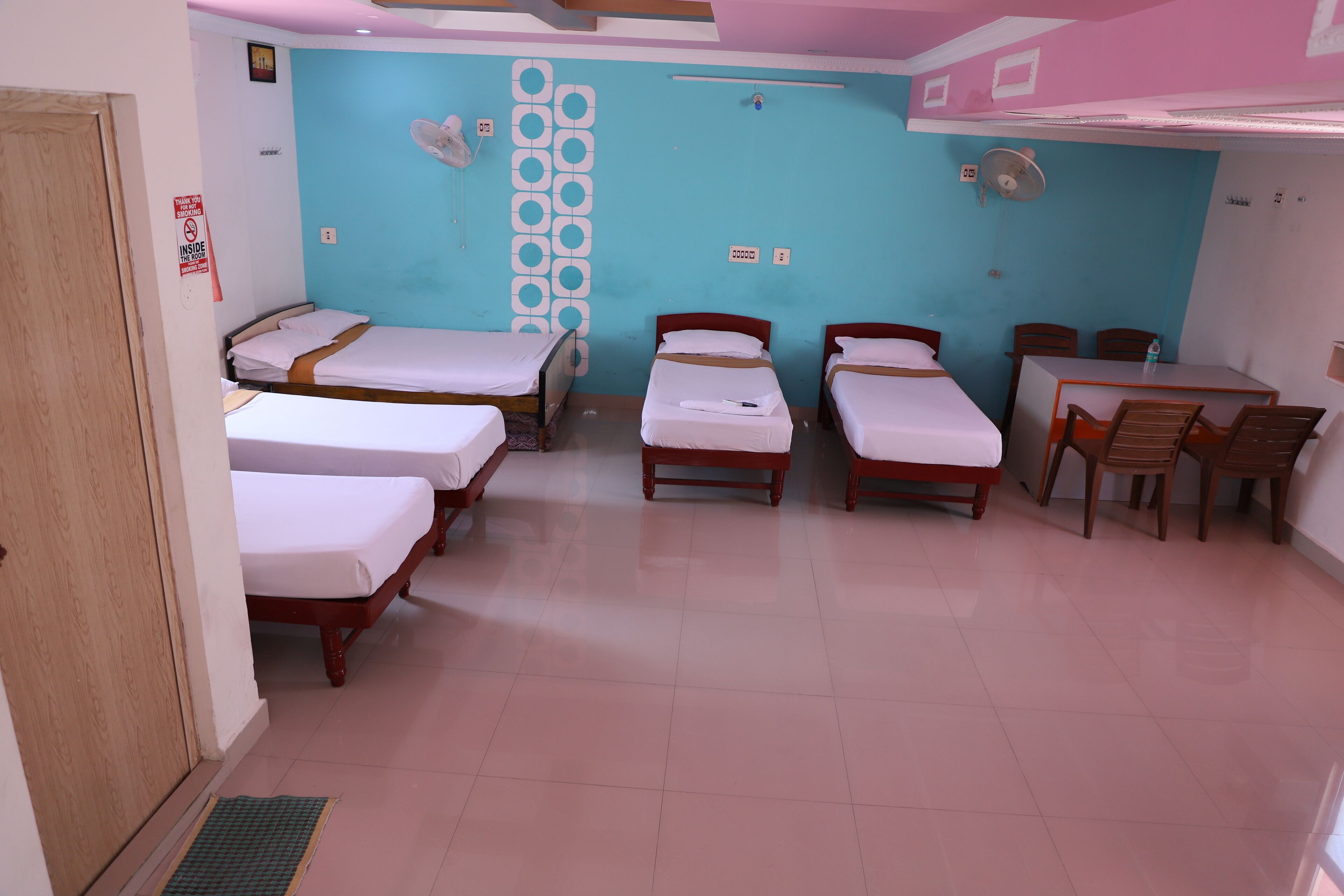 A hotel room with four single beds, a small table with chairs, and a wall-mounted fan.