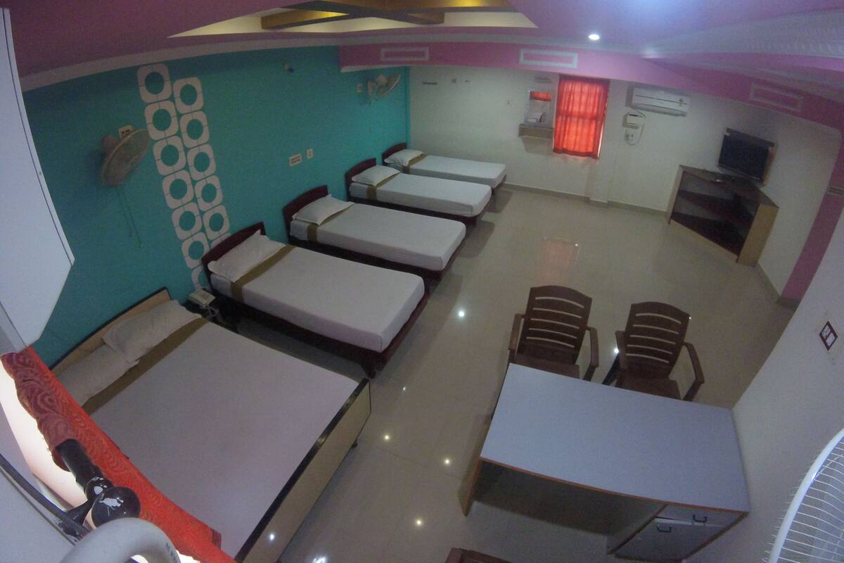 Quarto de hotel com quatro camas de solteiro, uma escrivaninha, duas cadeiras, televisão e uma pequena cozinha equipada.