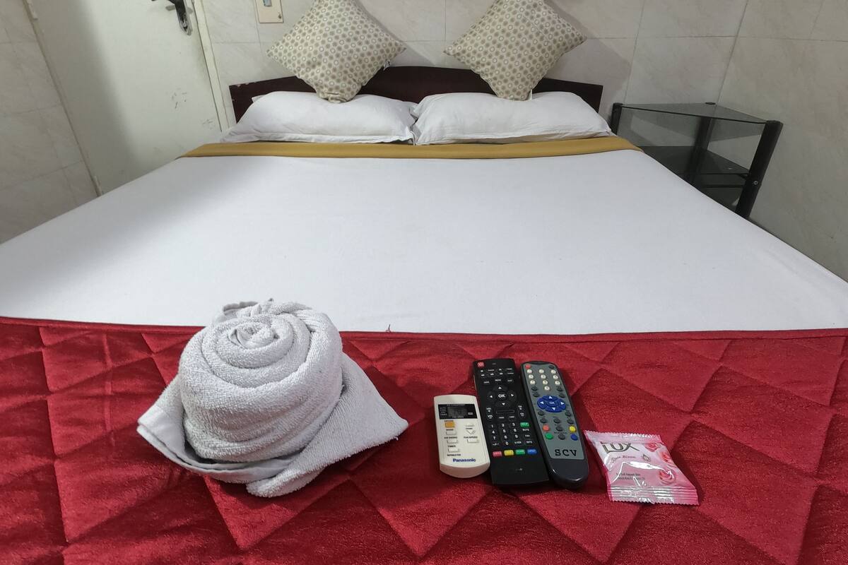 Uma cama bem arrumada com um edredom vermelho, dois travesseiros, uma toalha branca enrolada e três controles remotos na cama.