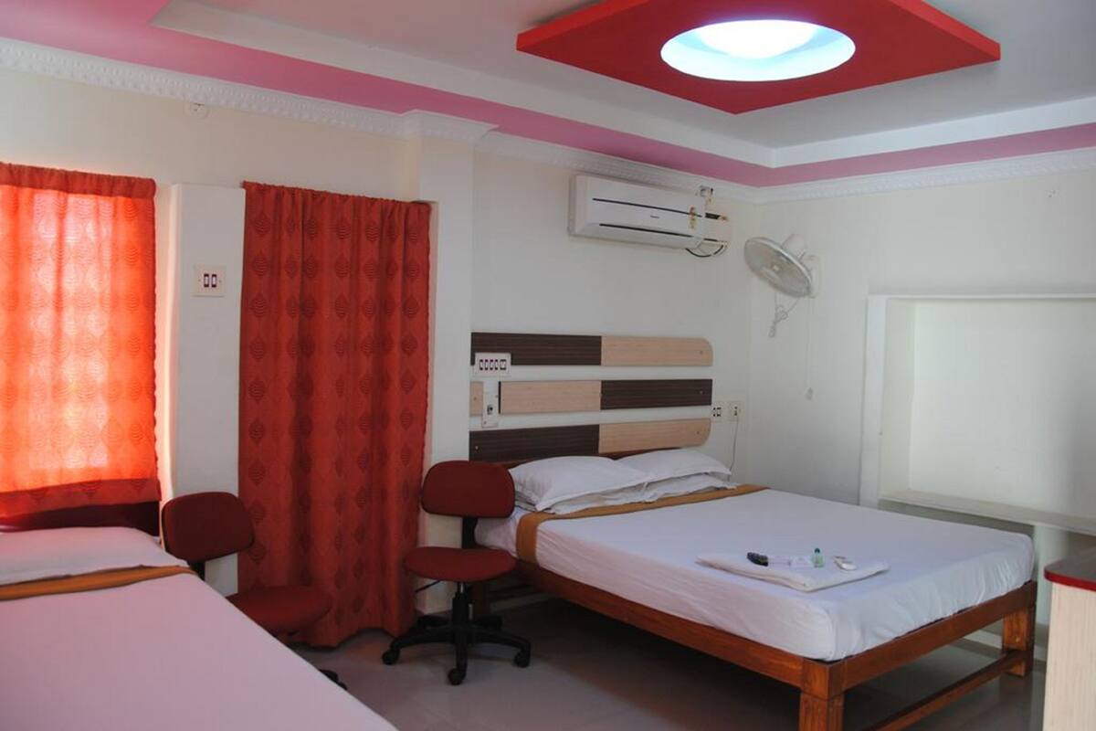 Quarto com duas camas, uma escrivaninha e uma cadeira. Há um ar condicionado na parede e uma luminária no teto.