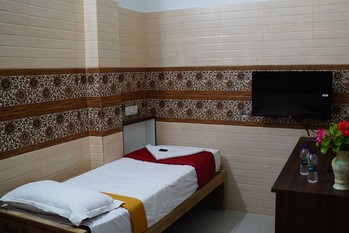 Quarto pequeno com uma cama de solteiro, cabeceira de madeira, televisão fixada na parede e mesa de cabeceira de madeira com um vaso de flores e garrafas de água.