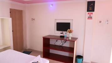 A hotel room with a bed, a TV on a stand, a small table with a vase and a remote control, a door, and a trash bin.