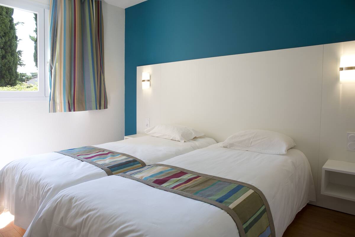 A hotel room with two beds, a window with curtains, and a wall-mounted light fixture.