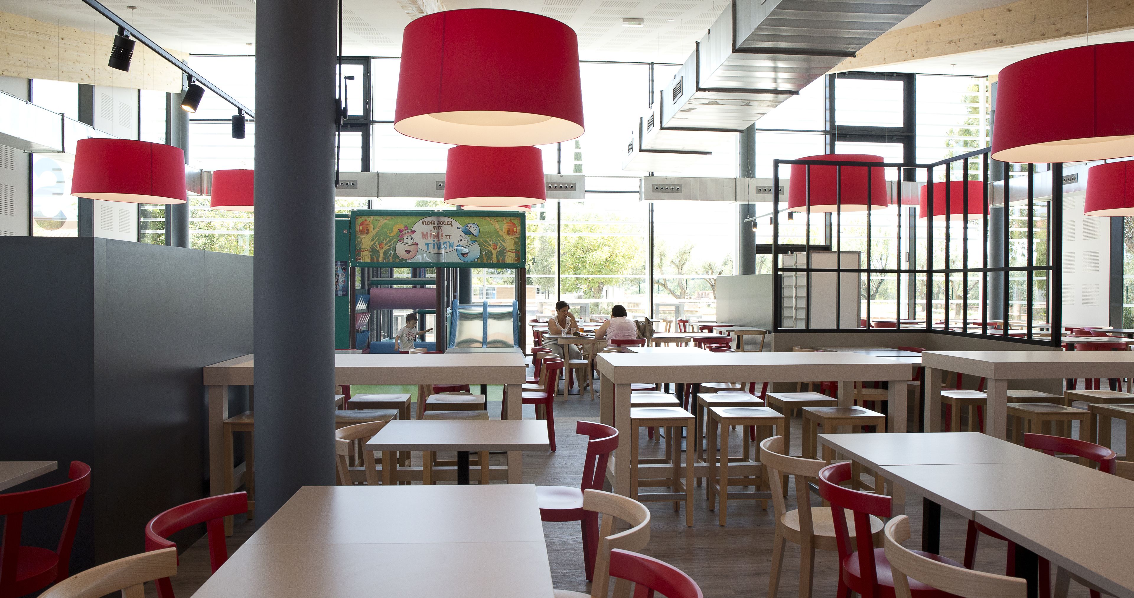 A restaurant interior with multiple tables and chairs, large windows, and red hanging lamps.