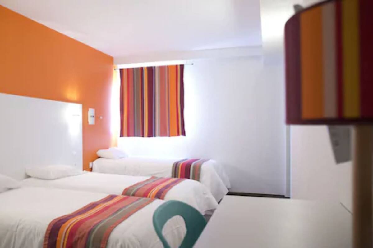 A hotel room with two beds, white bedding, a striped curtain, and orange walls.