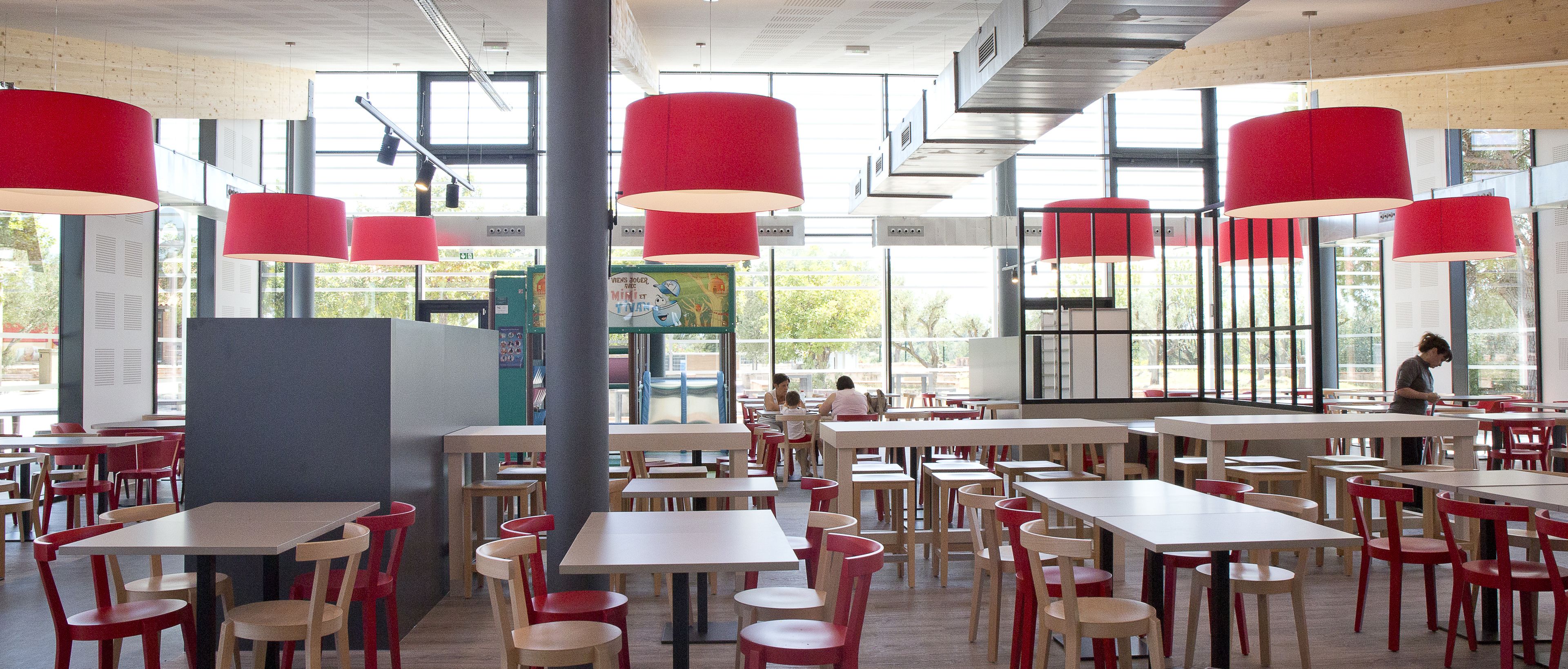 A spacious cafeteria with high tables and chairs, large windows, and red pendant lights.