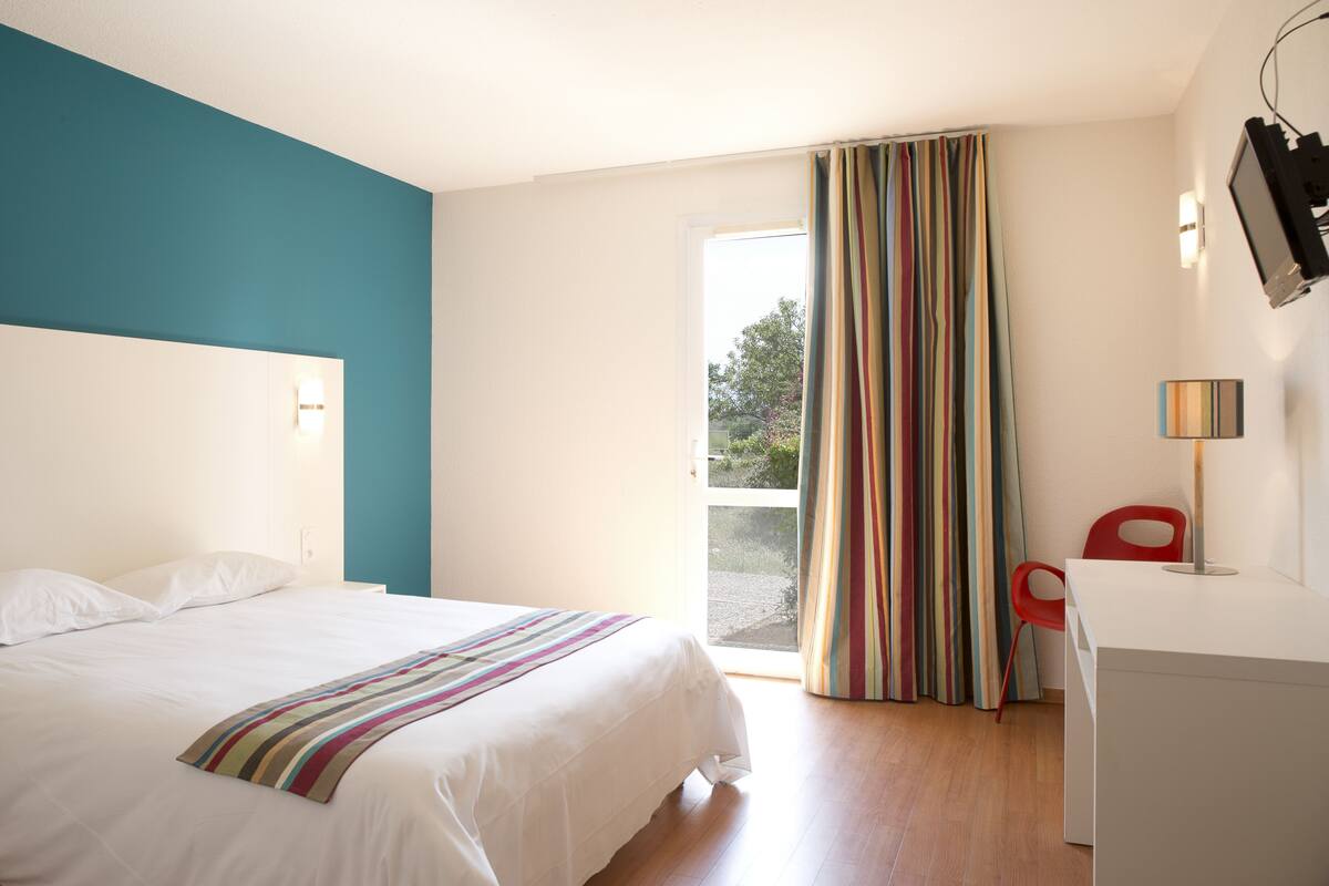 A hotel room with a bed, a desk, a chair, a TV, and a window with curtains.