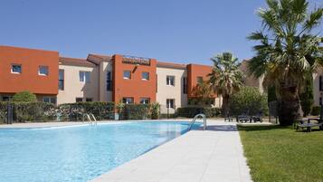 Una piscina con borde de baldosas, rodeada de palmeras y un edificio con múltiples ventanas y balcones.