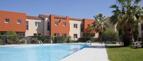 Una piscina con borde de baldosas, rodeada de palmeras y un edificio con múltiples ventanas y balcones.