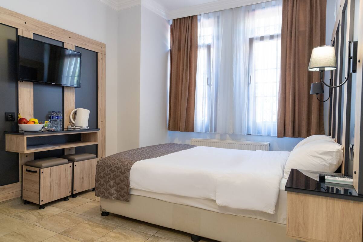 A hotel room with a bed, a TV mounted on the wall, a desk with a computer, and a window with curtains.
