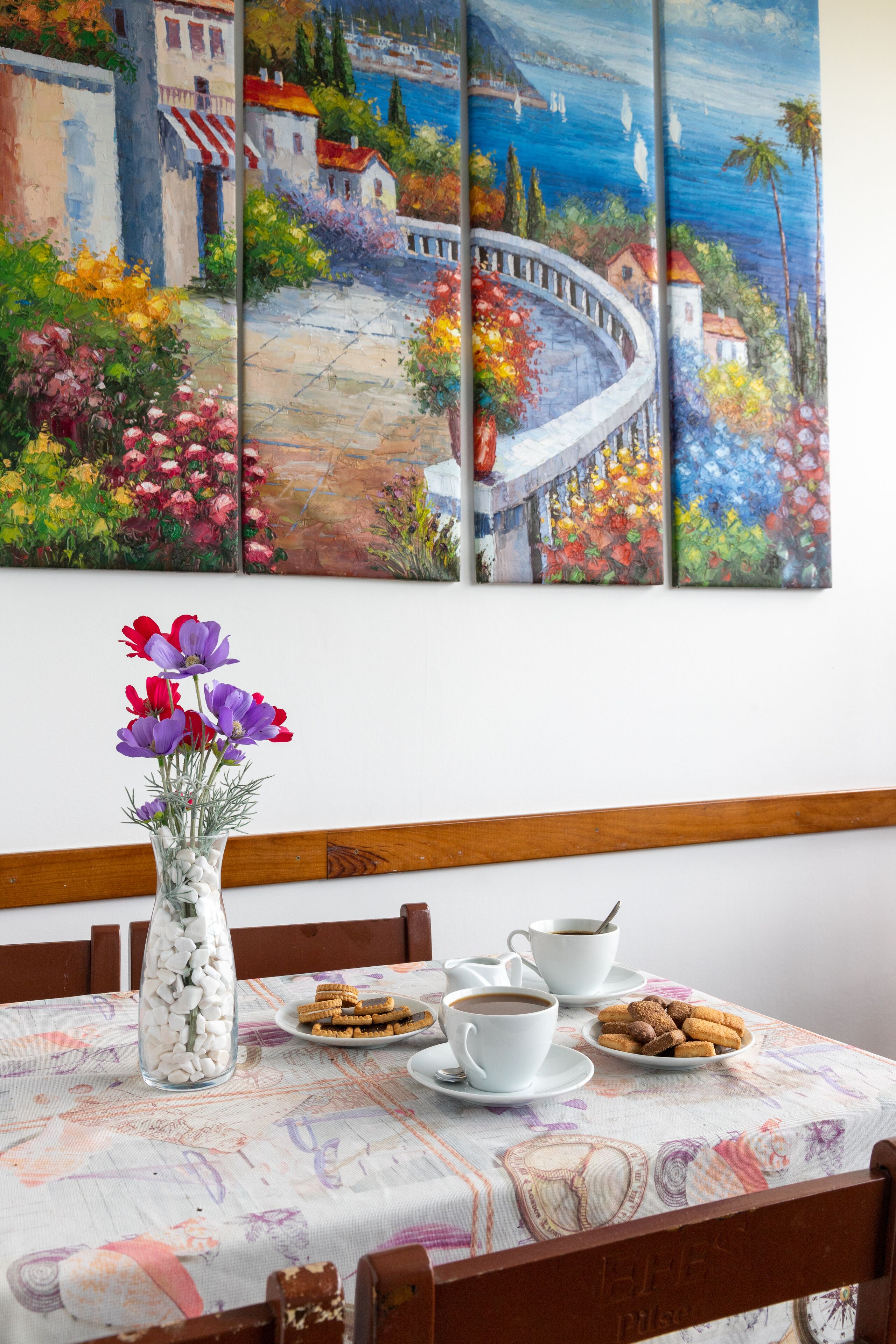 A dining table with a painting of a coastal scene, a vase with flowers, and a plate of cookies.