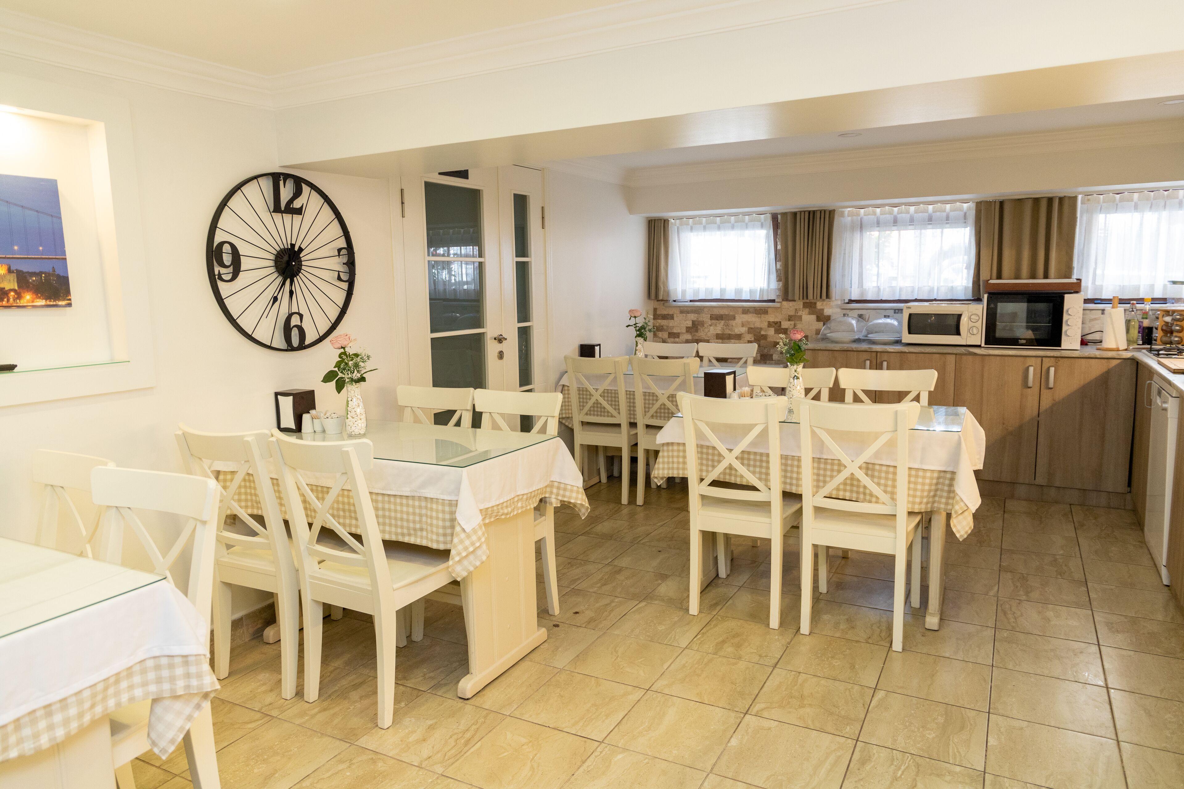 A dining area with white chairs and tables, a large wall clock, and a kitchen with a microwave and oven.