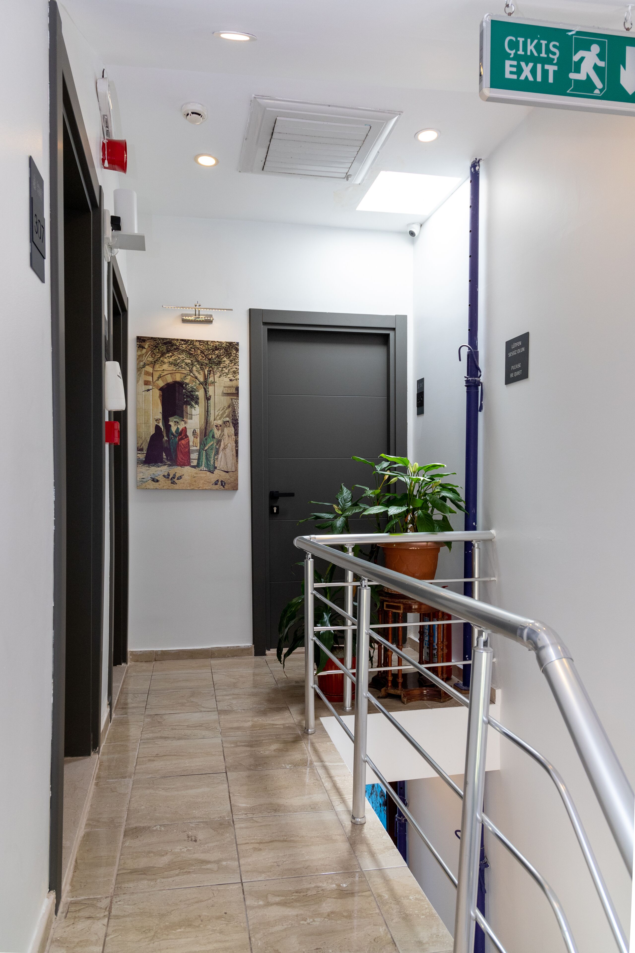 A hallway with a tiled floor, a door, a green EXIT sign, a potted plant, and a painting on the wall.