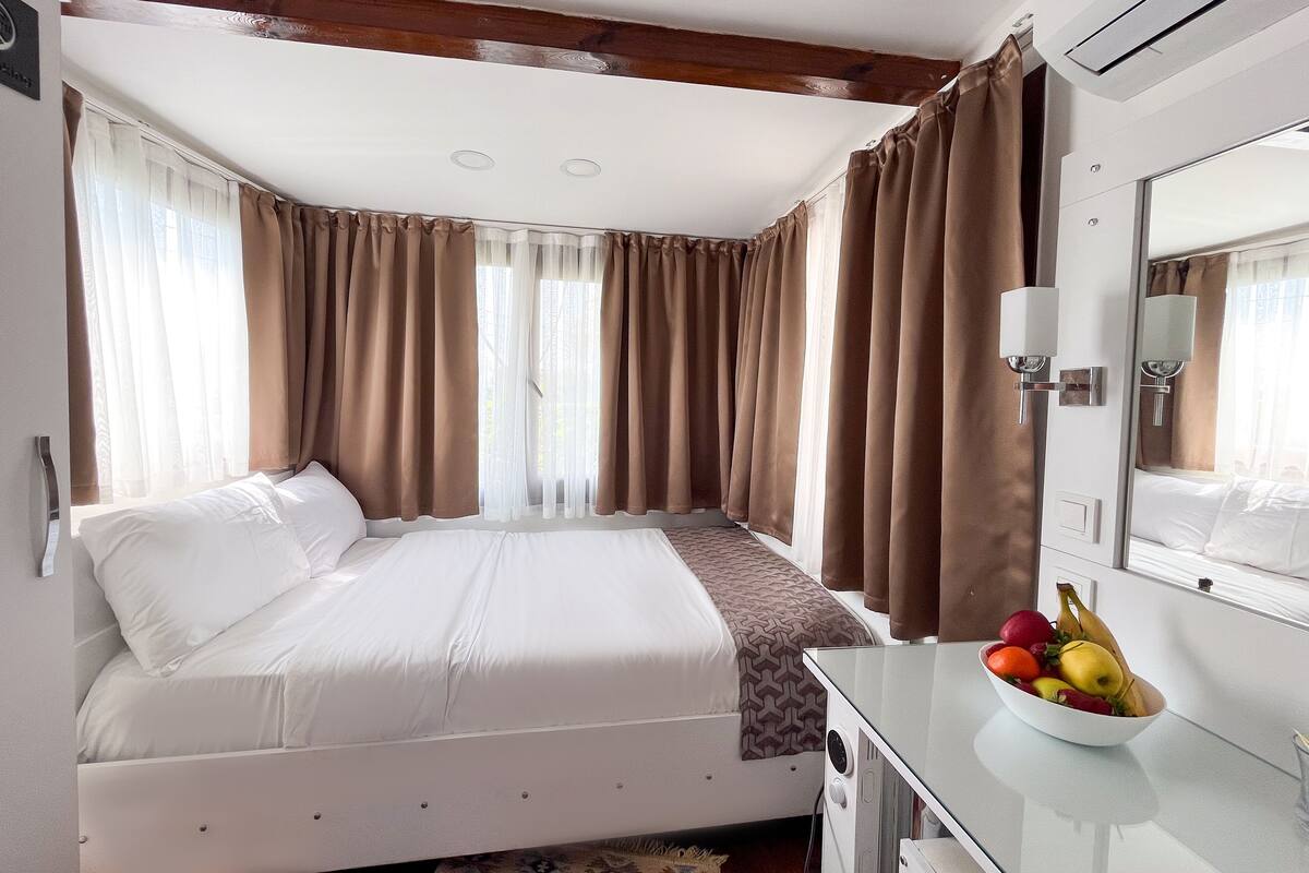 A hotel room with a bed, a bedside table with a fruit bowl, a mirror, and curtains.