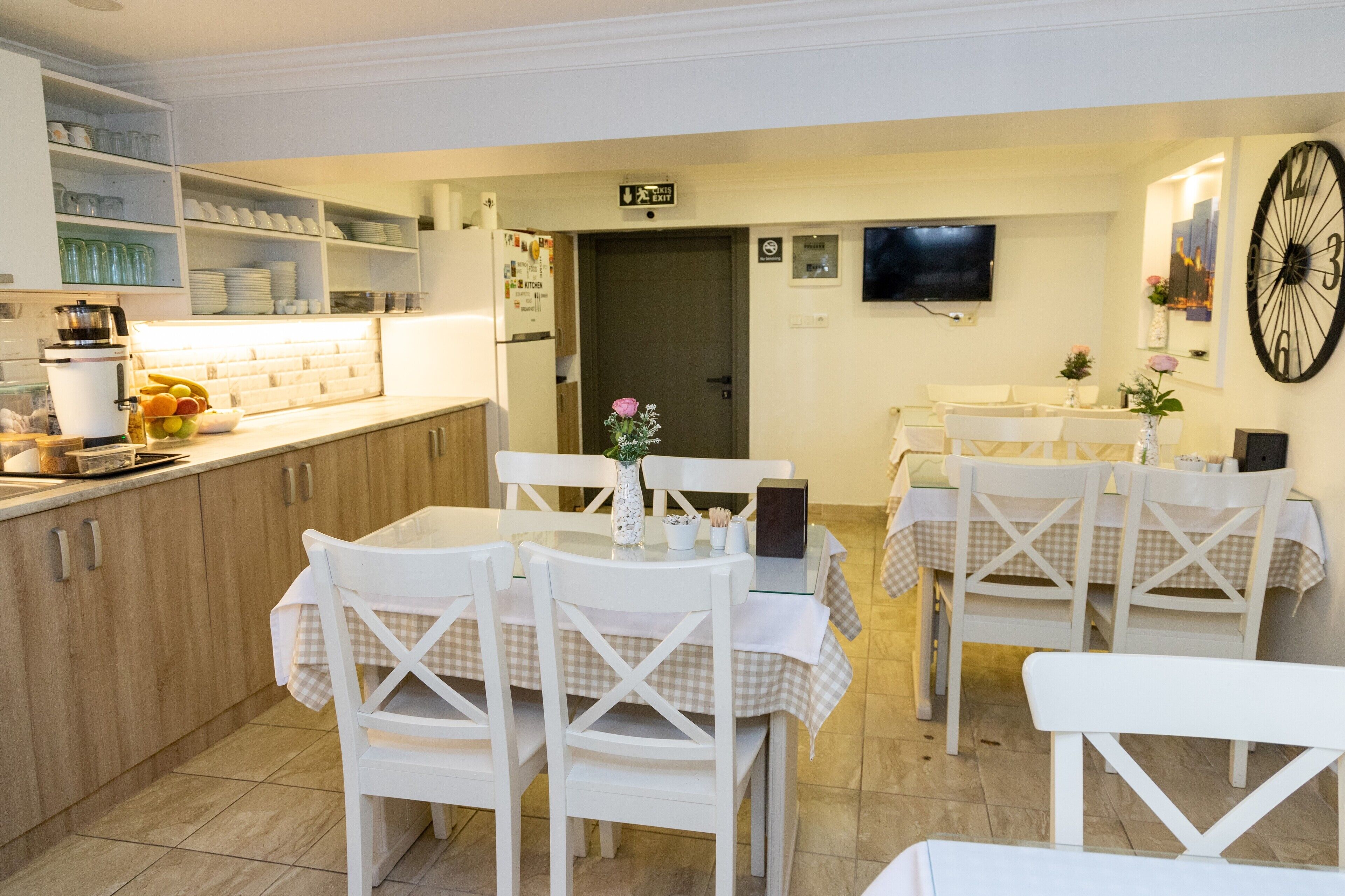 A dining area with white chairs and tables, a kitchen with shelves, and a TV mounted on the wall.