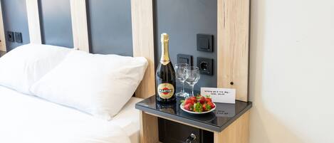 A hotel room with a bed, a minibar with a bottle of Martini champagne, two glasses, and a plate of strawberries.