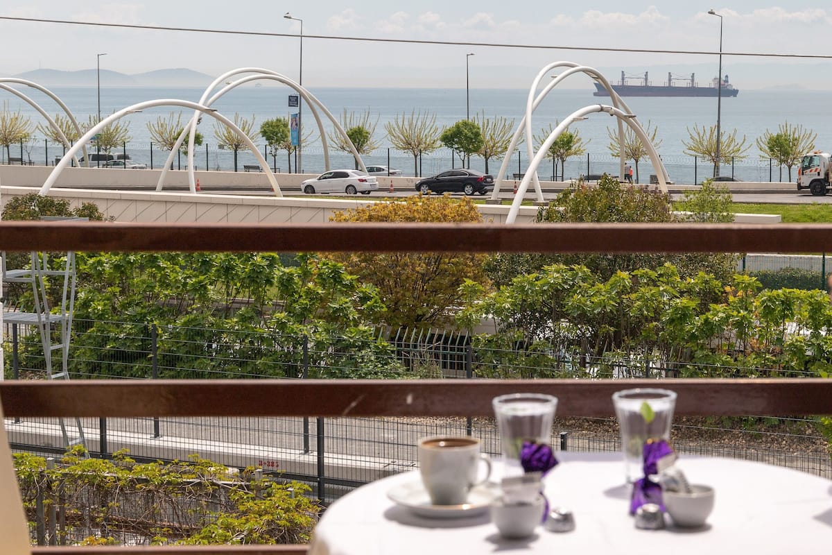 A table with a cup and saucer, a glass, and a small bowl, set against a backdrop of a coastal view with a bridge and ships in the distance.