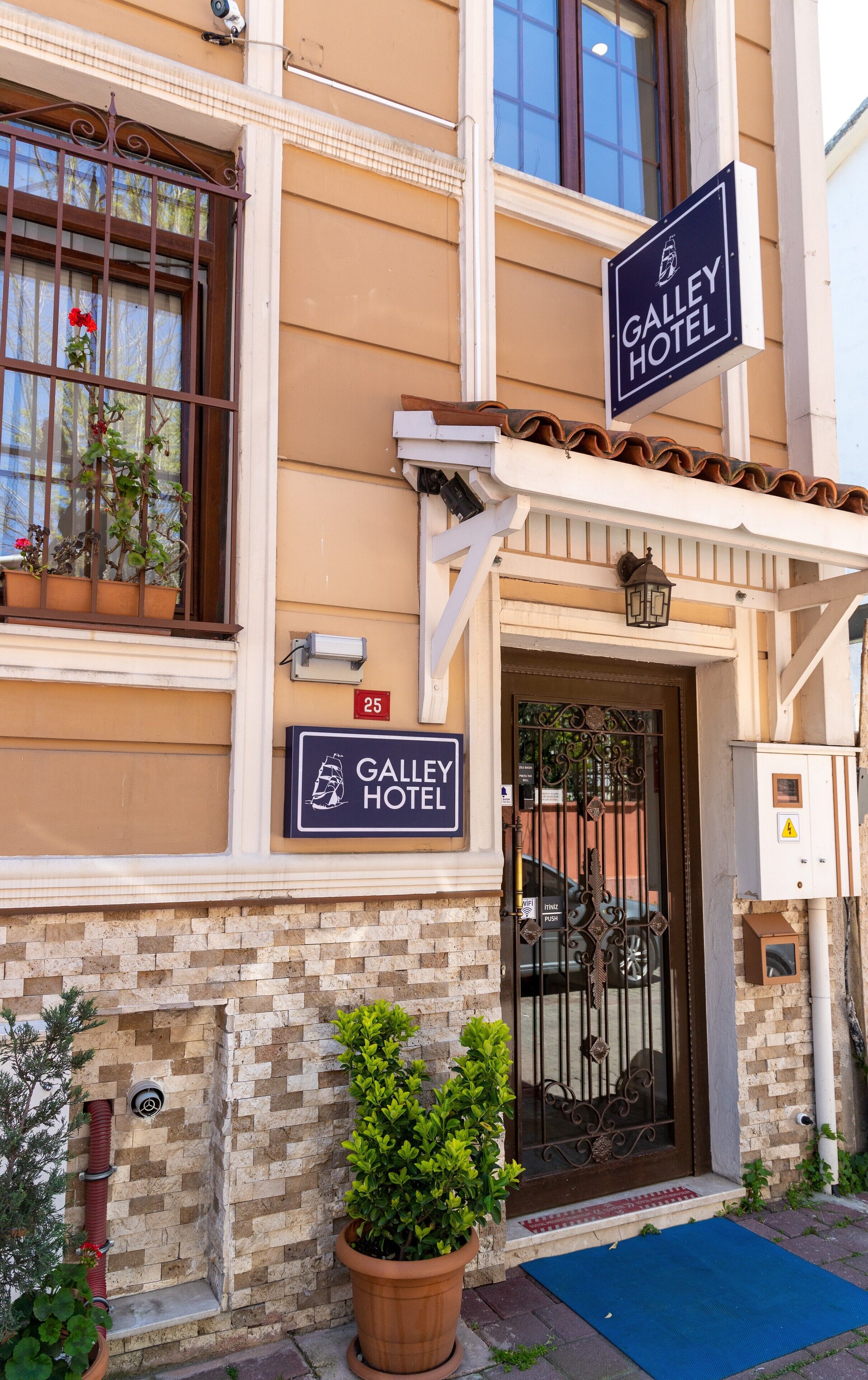 L’entrée d’un hôtel, avec un portail en fer forgé, un paillasson bleu et un pot de fleurs.