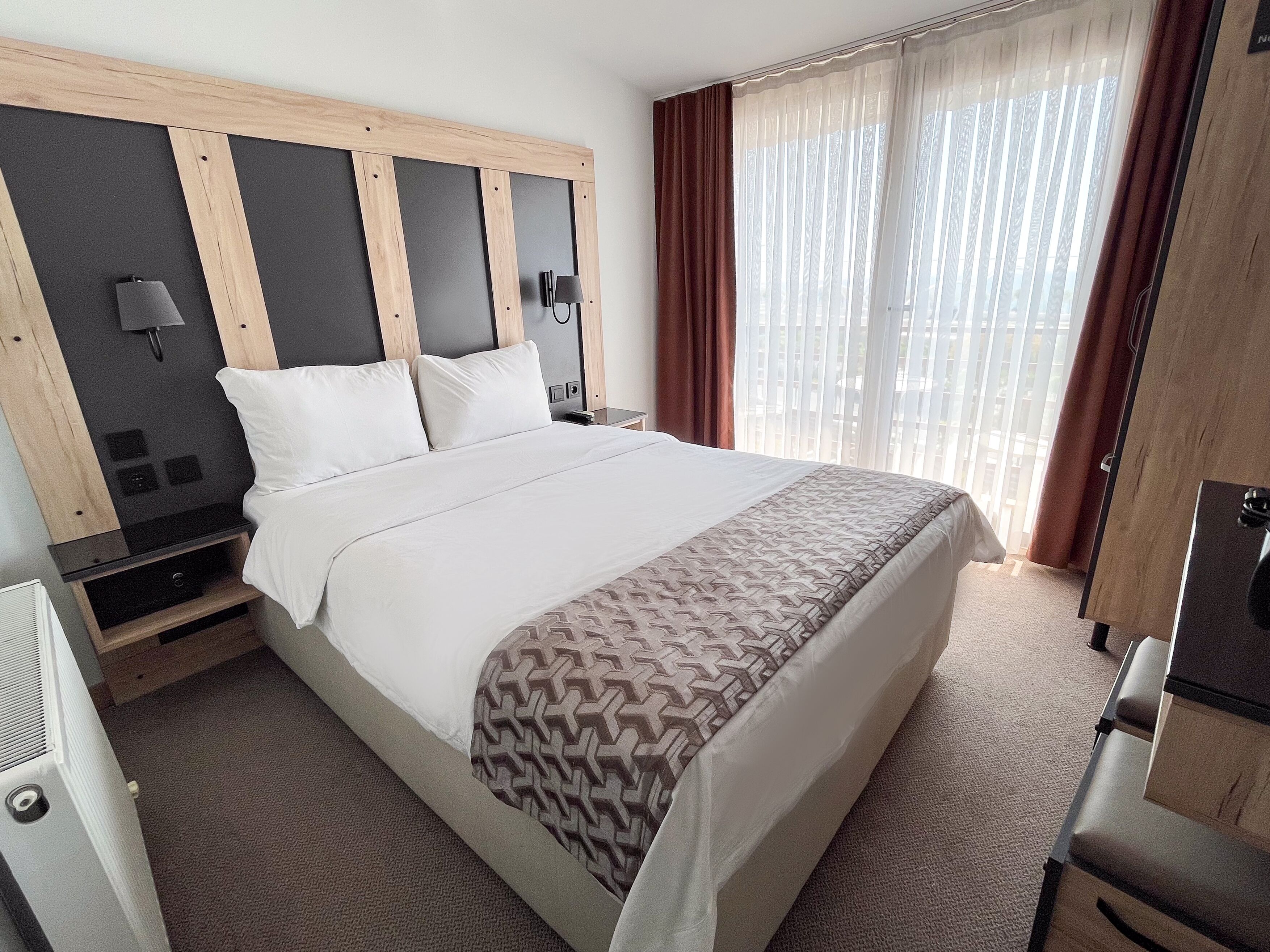 A hotel room with a large bed, a wooden headboard, a bedside table with a lamp, and a window with curtains.