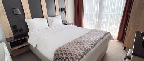 A hotel room with a large bed, a wooden headboard, a bedside table with a lamp, and a window with curtains.