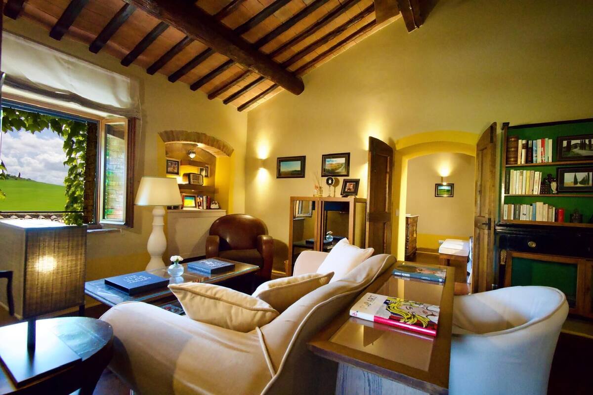 A cozy living room with a sofa, armchair, and a coffee table. There is a bookshelf filled with books and a lamp on a side table. A window with a view of a green landscape is visible.