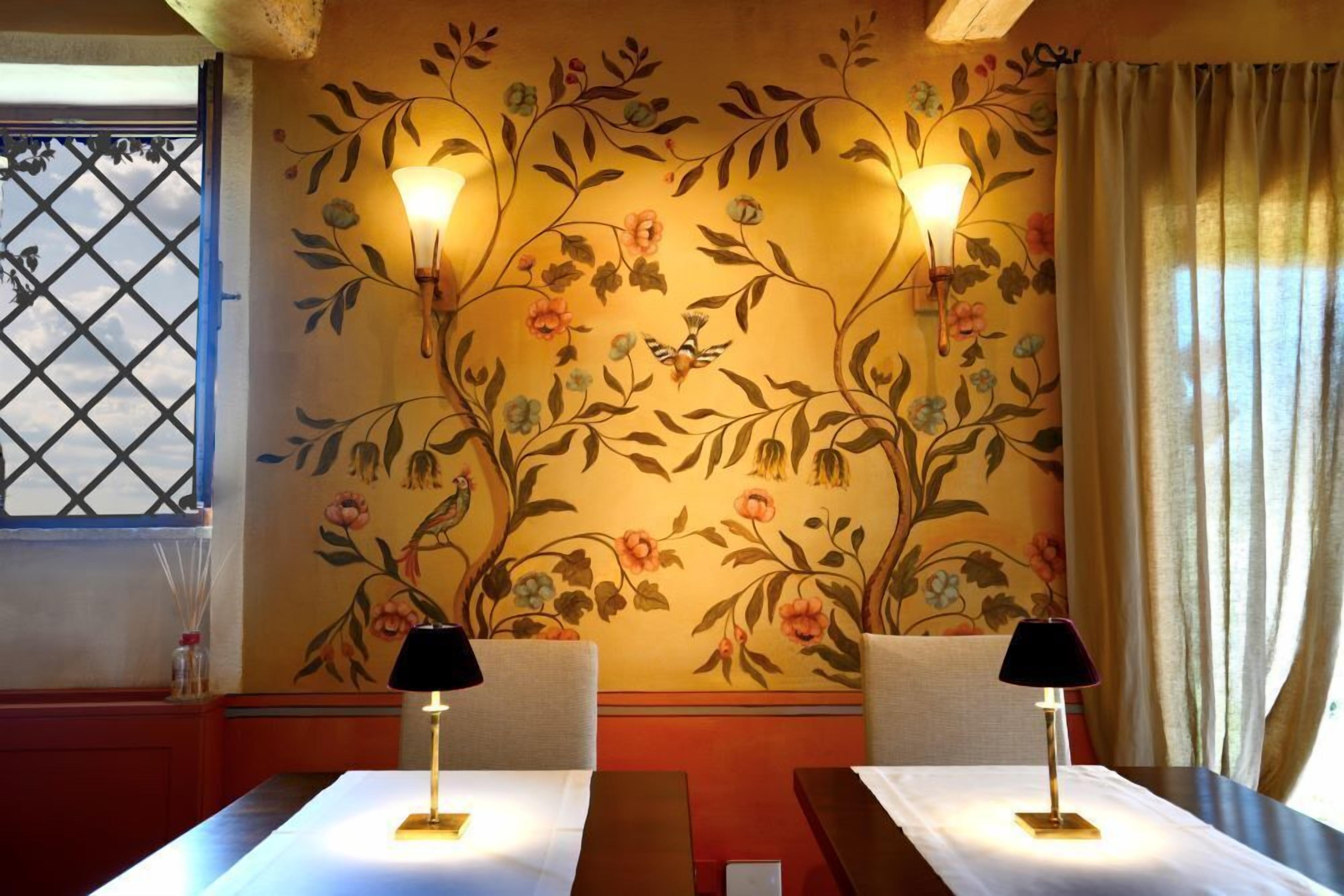 A dining area with two tables, each with a lamp and a white tablecloth. The walls are decorated with a floral mural.