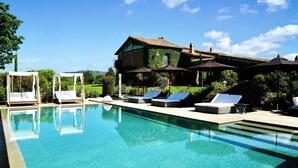 Una piscina con tumbonas y sombrillas, rodeada de abundante vegetación y una casa con techo de tejas.