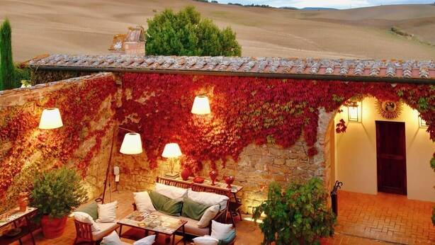 Un espacio al aire libre acogedor con muros de piedra y follaje rojo, que incluye una mesa y sillas, y una puerta que da a un acceso cubierto.