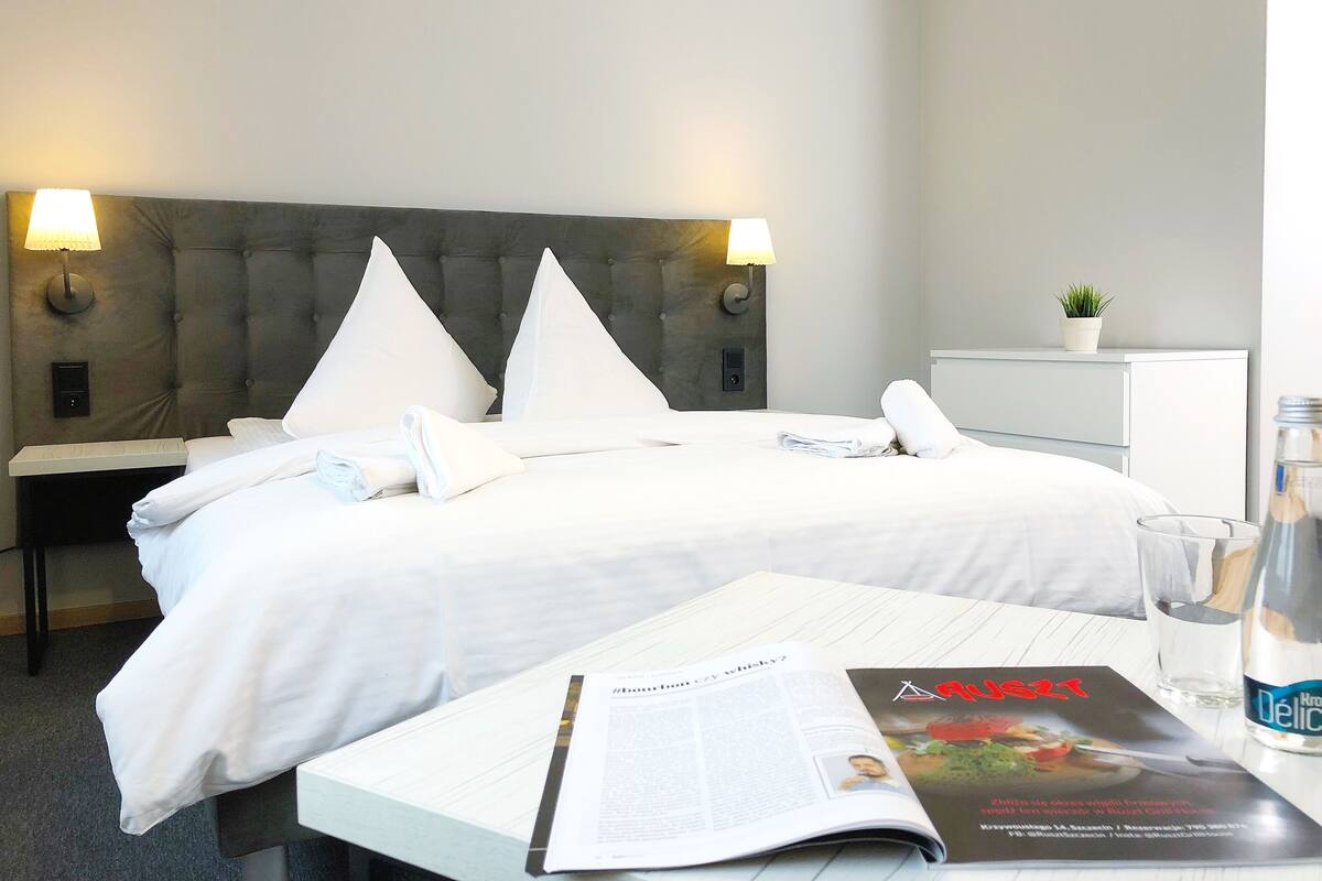 Una cama bien hecha con sábanas blancas, dos lámparas de noche, una pequeña planta en maceta en una mesita de noche y una revista sobre una mesa.