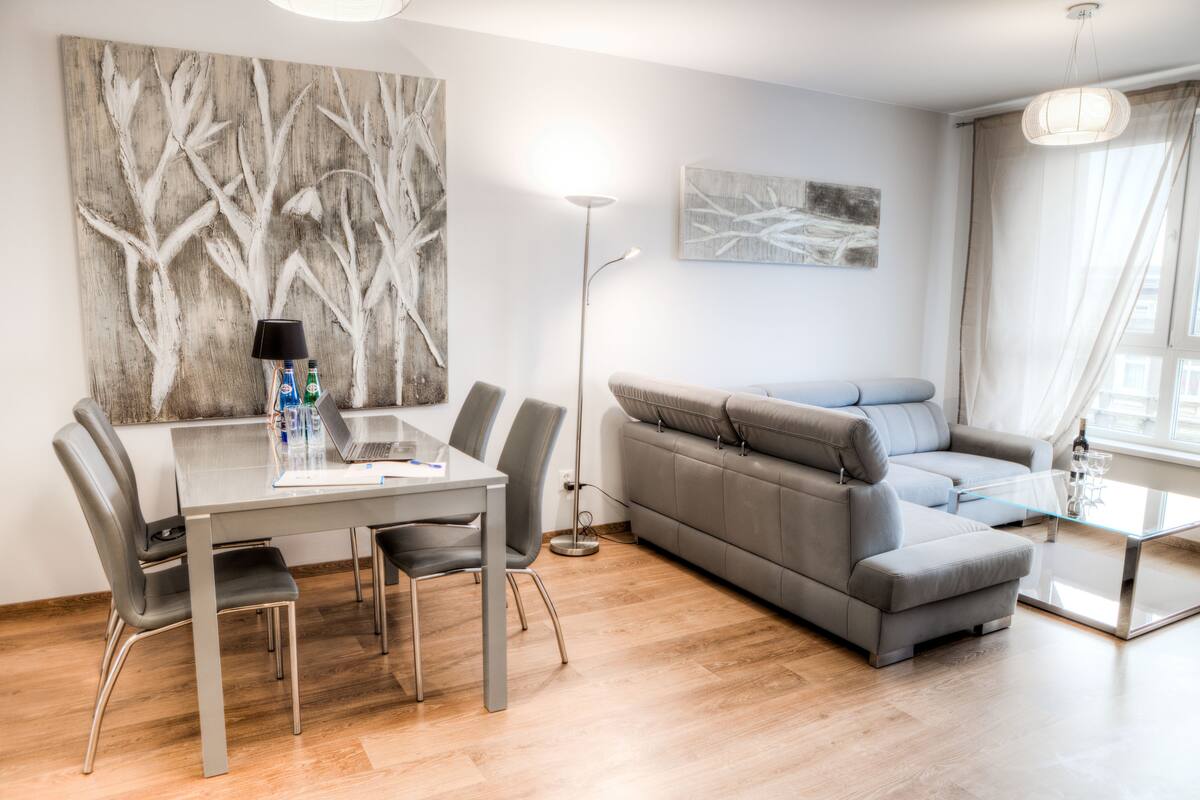 Una sala moderna con un sofá gris, una mesa de comedor con sillas, una lámpara de pie y obras de arte en las paredes.
