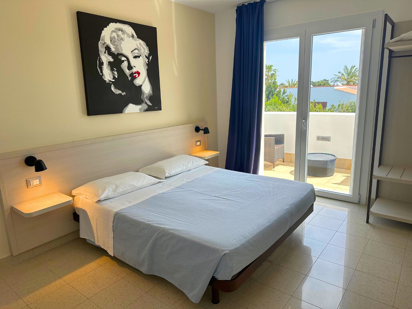 A bedroom with a bed, two bedside tables, a wall-mounted lamp, a large window with a view of the outdoors, and a sliding glass door leading to a balcony.