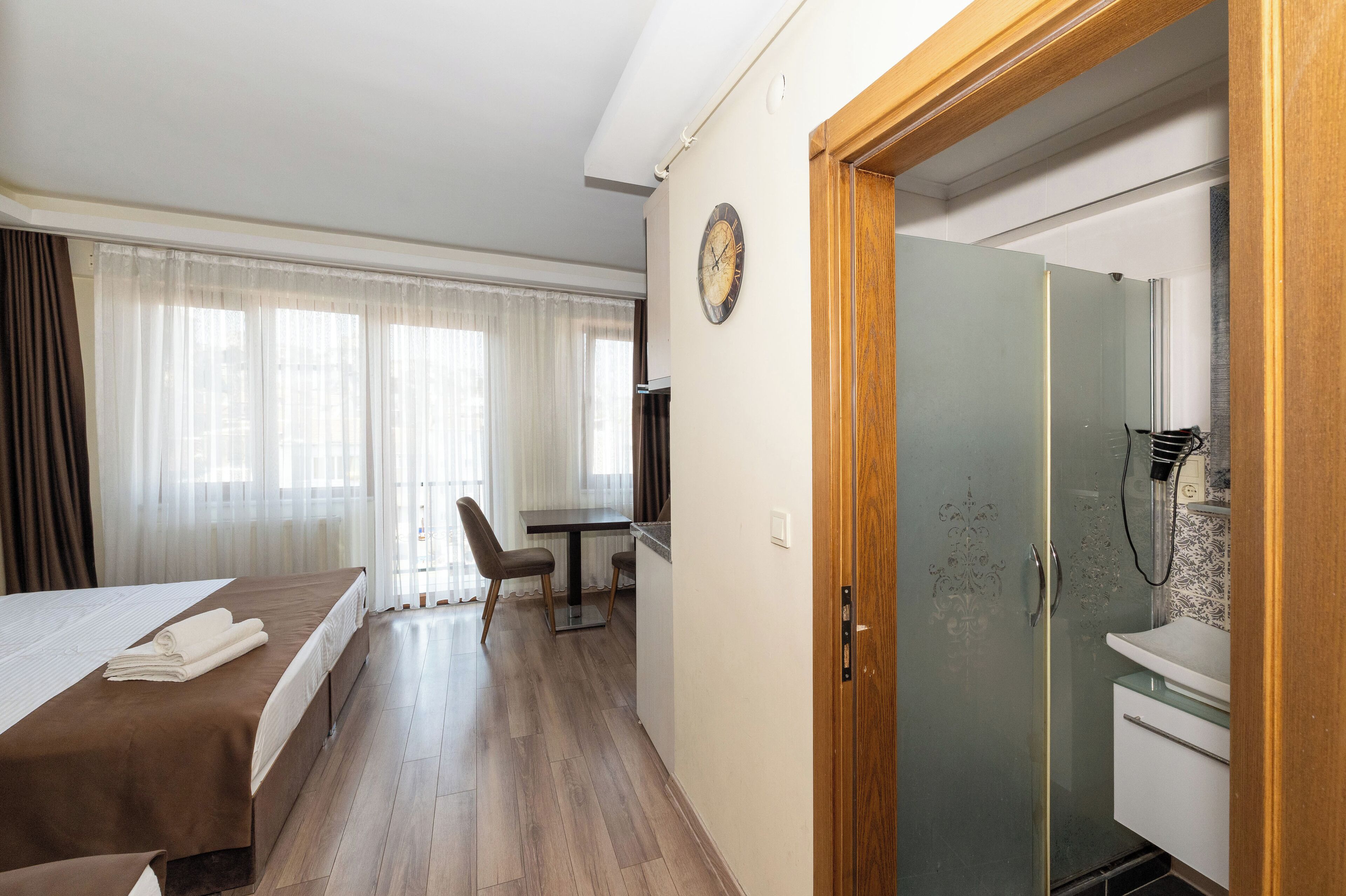 A hotel room with a bed, a desk, a chair, a clock, and a shower area.