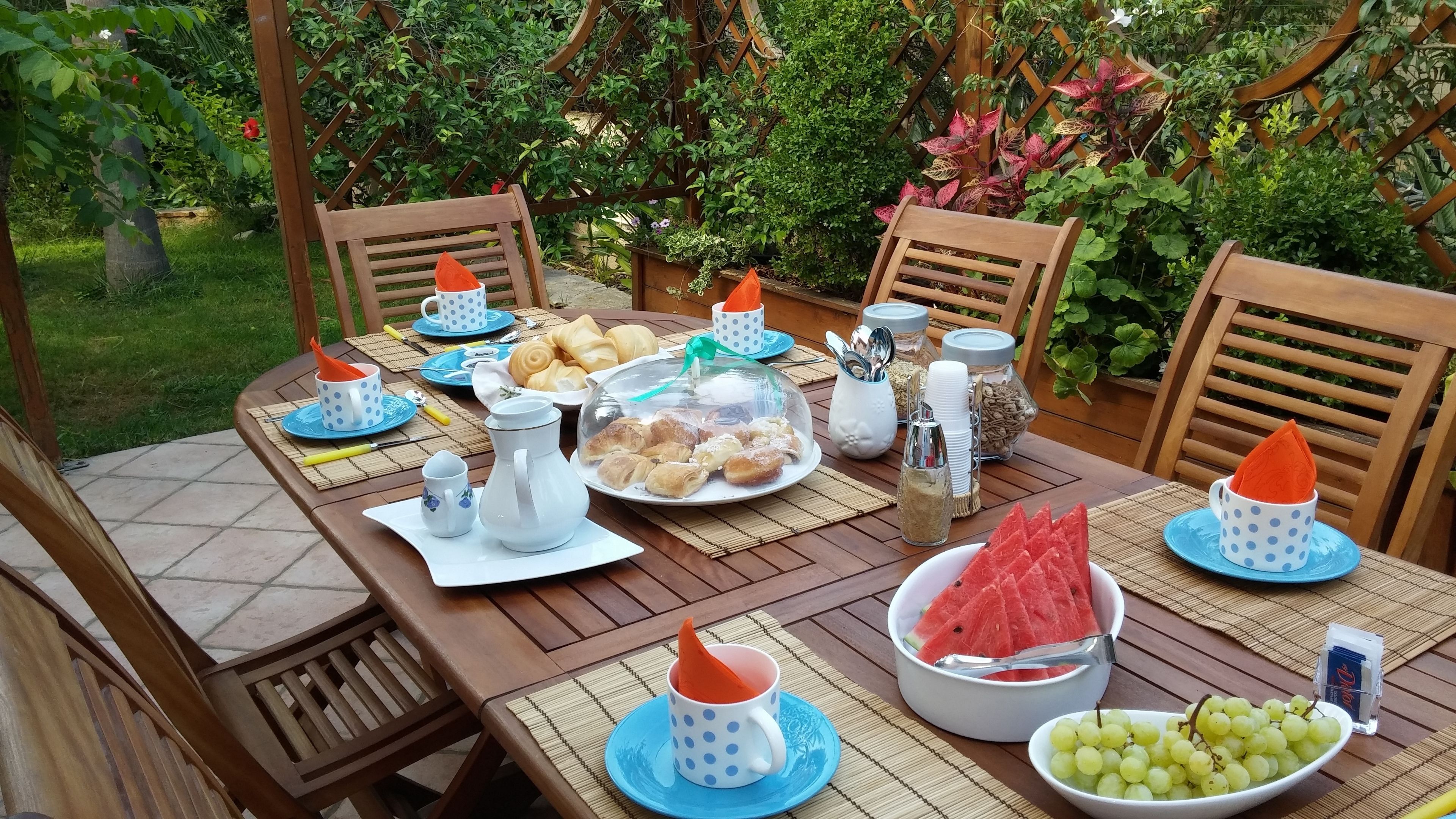 Una mesa de madera preparada para una comida, con platos, tazas y una jarra, rodeada de sillas en un jardín.