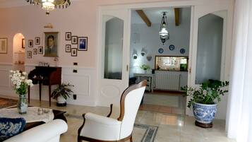 Amplia sala de estar con un sillón blanco, una mesa de centro y un jarrón de flores. Hay un candelabro colgado del techo y una puerta que da a una zona de cocina.