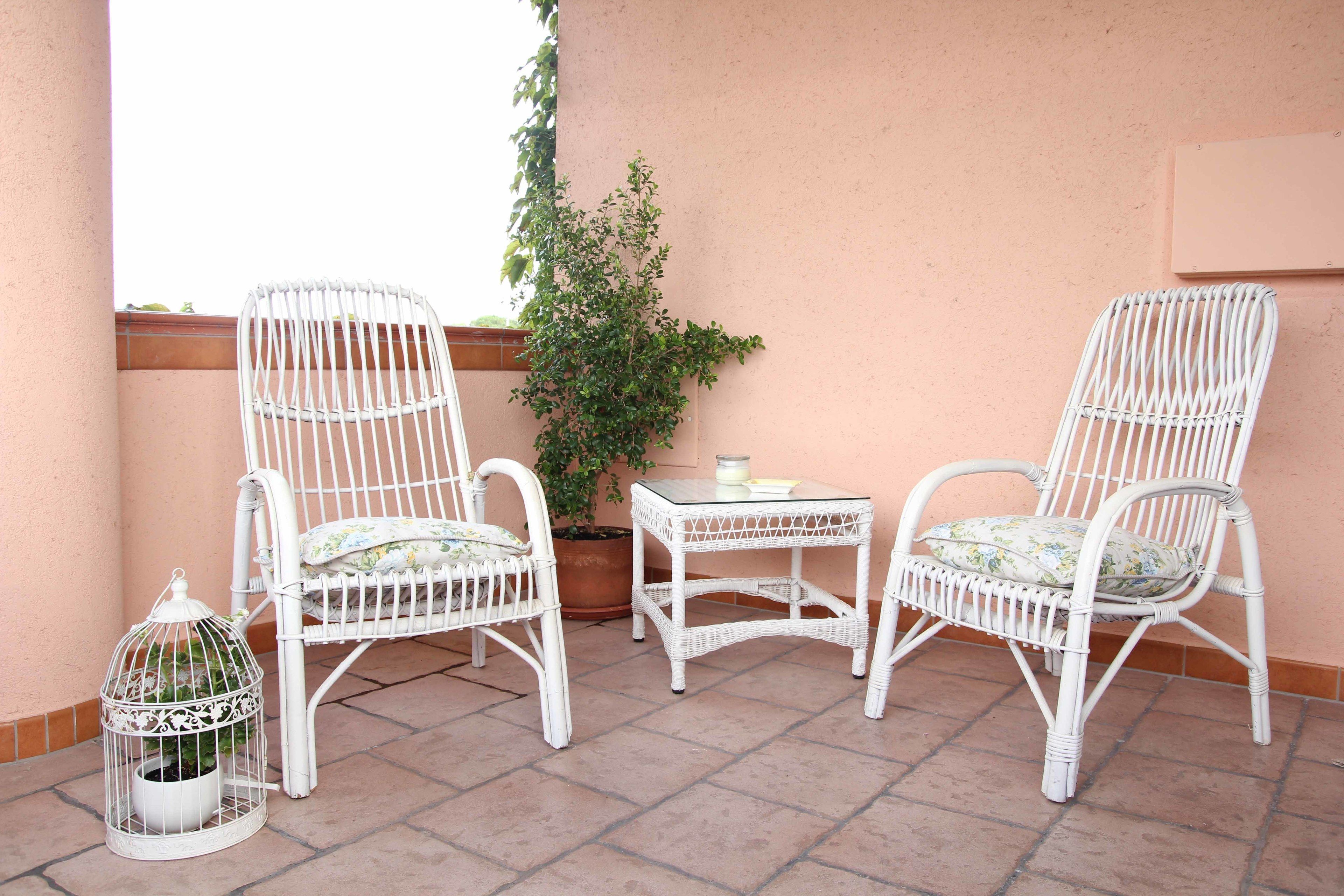 Deux chaises en osier blanc avec des coussins à motifs floraux, une petite table et une plante en pot sur une terrasse carrelée.