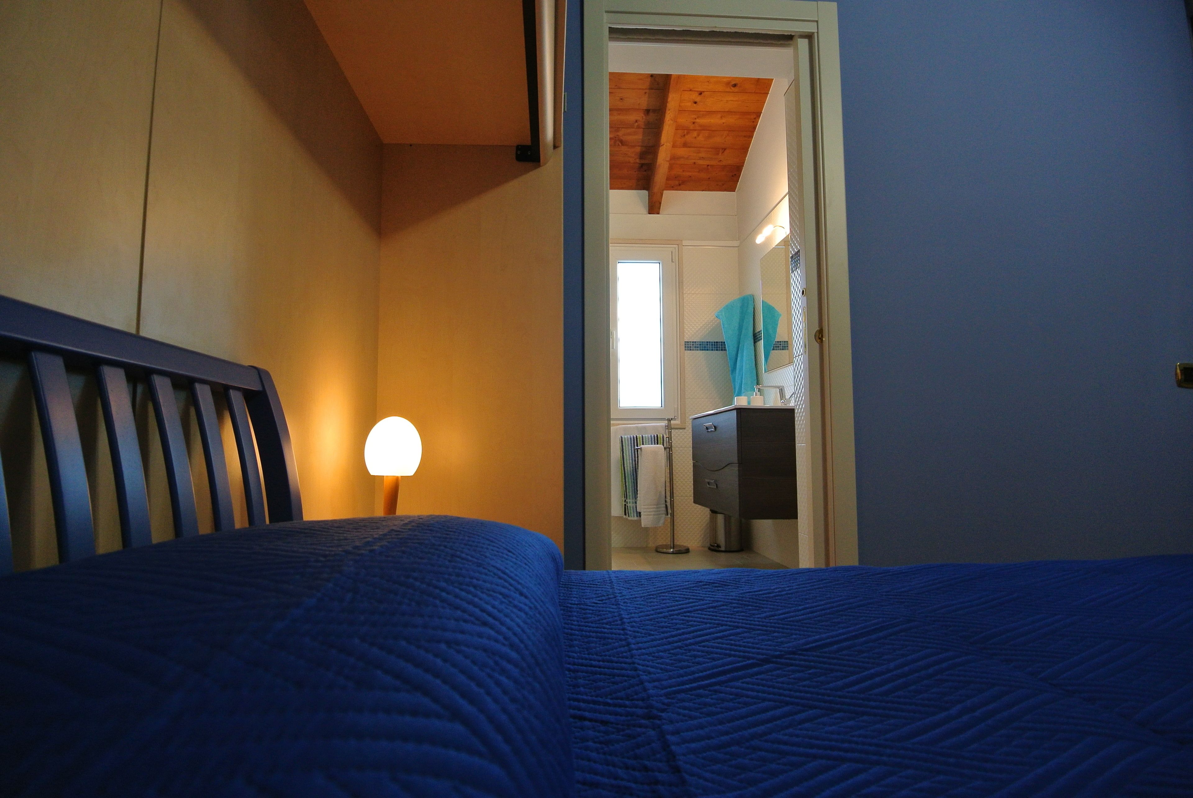 Un dormitorio con cama, mesita de noche, lámpara y vistas a un baño con toallero y espejo.