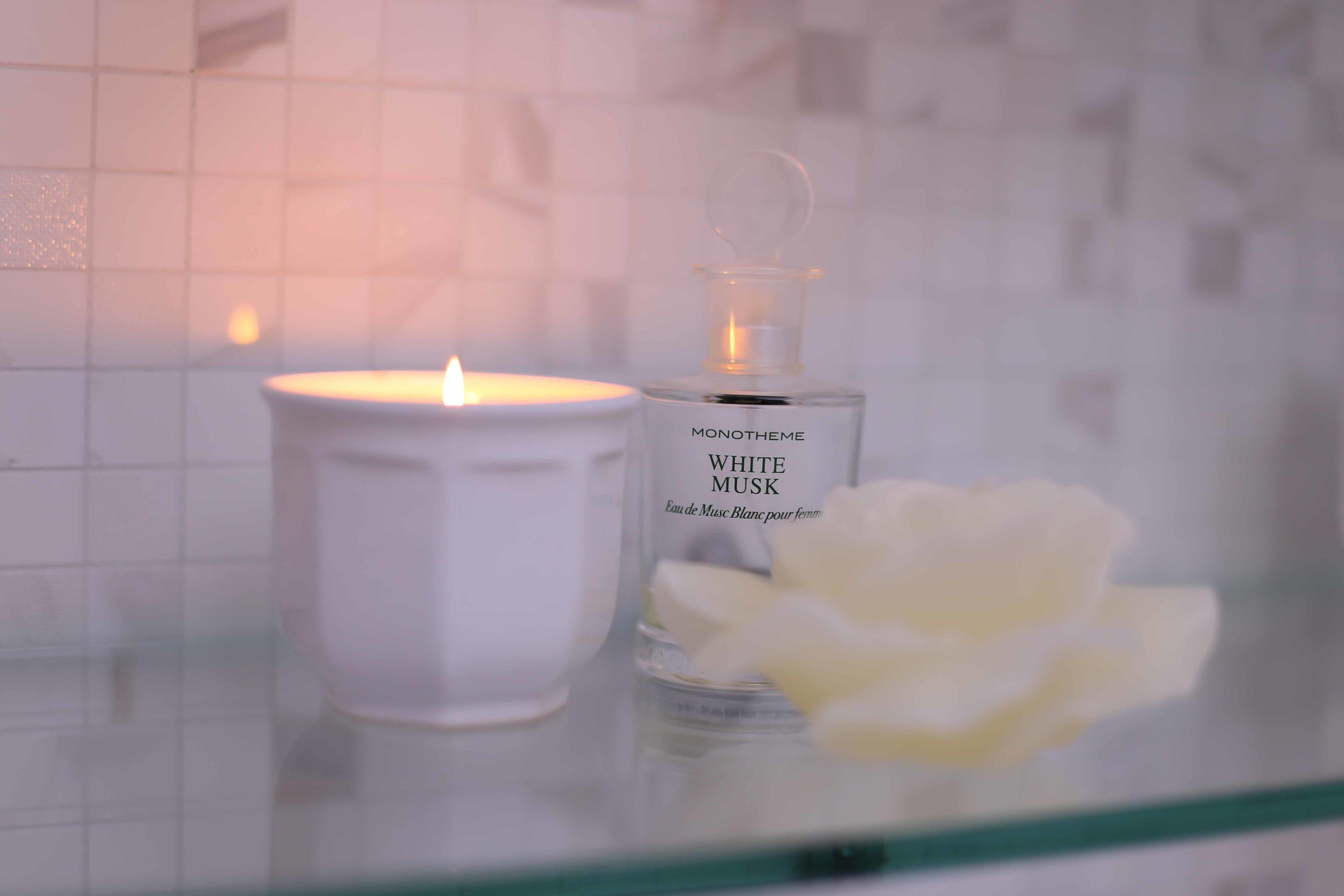 Una vela blanca y un frasco de colonia Monotheme White Musk sobre una superficie de vidrio.